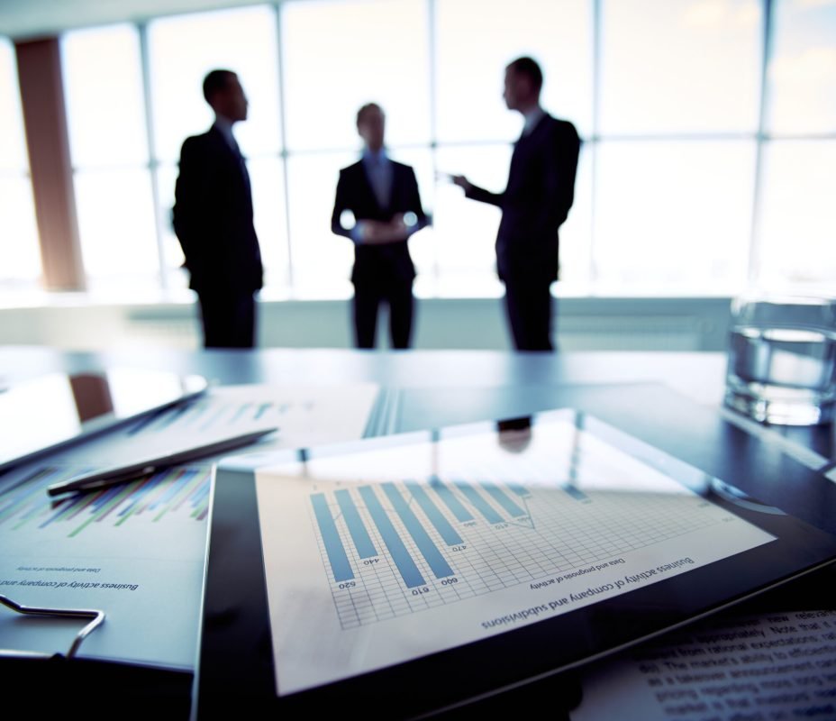 Close-shot of a tablet computer displaying financial data, three businessmen standing in the background